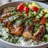 Sliced, juicy chimichurri chicken rests atop a bed of fluffy rice with diced cucumbers, red onions, cherry tomatoes, and avocado in a vibrant bowl.